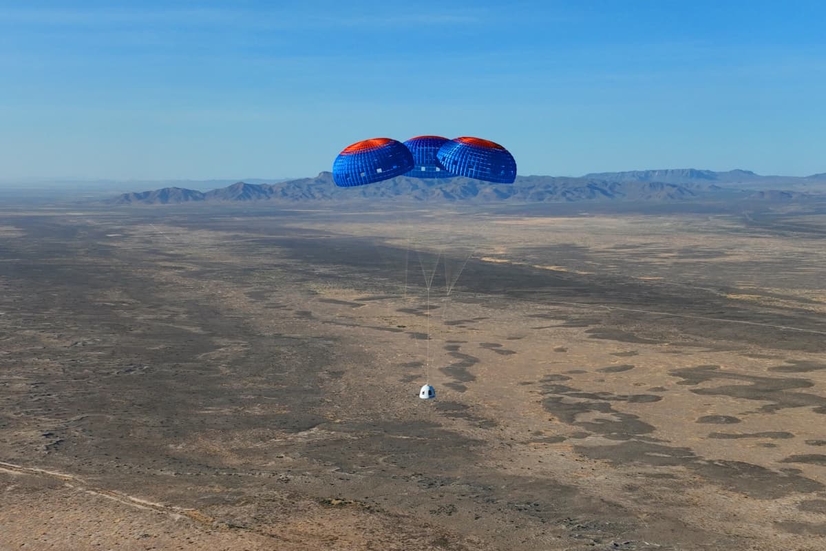 The crew capsule descends under its main blue and orange  parachutes; the capsule is small against the vast desert with the mountains in the distance.