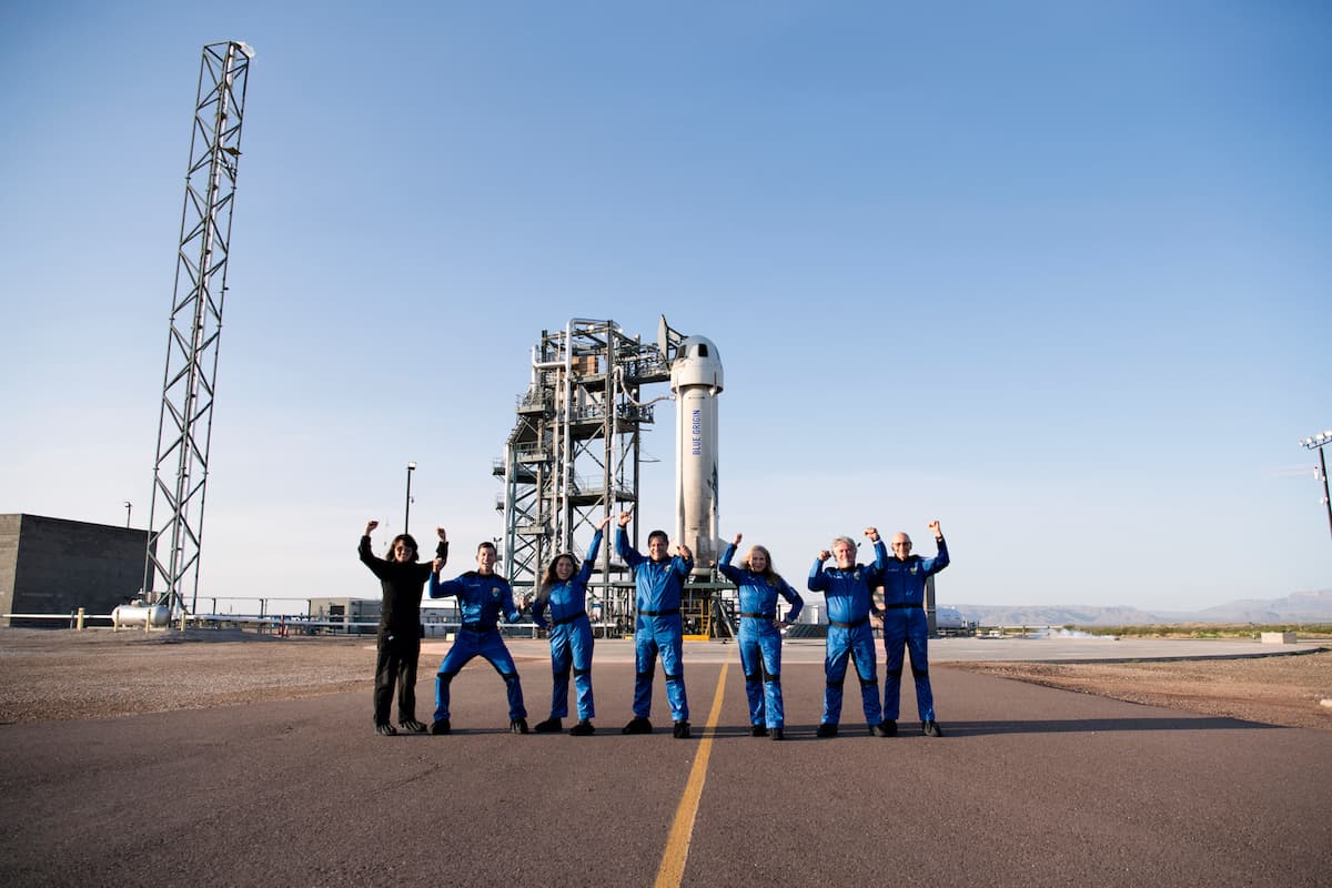 The crew comprising a mix of genders and ethnicities pose with their arms raised in excitement prior to their flight; they're standing on the ring road encircling the vehicle on the launch tower.