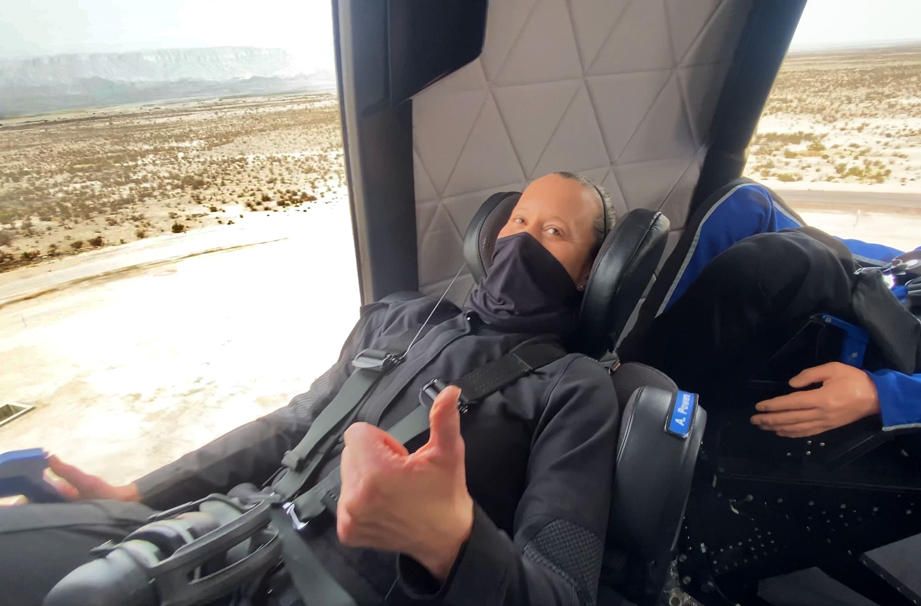 Audrey gives a thumbs up and wears a black flight suit and face mask while harnessed into a crew capsule seat during astronaut rehearsals. The desert can be seen in the background.