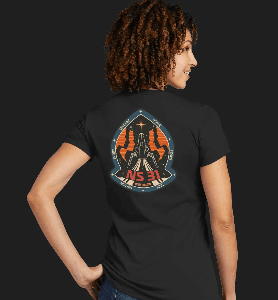 A person facing away from the camera wears a black T-shirt with the NS-31 mission patch on the back.