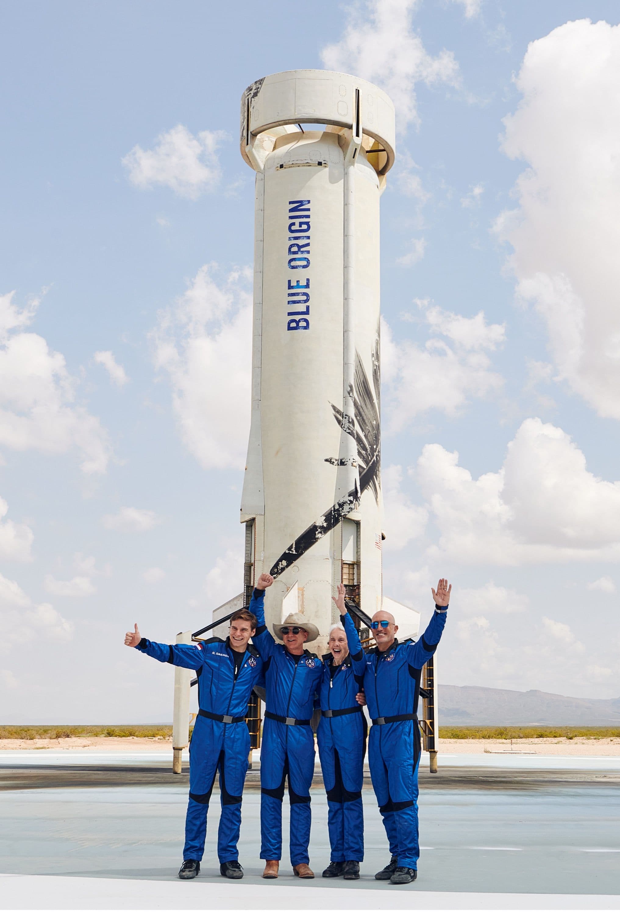 The crew stands shoulder-to-shoulder in front of the booster on the landing pad grinning with their arms raised in celebration.