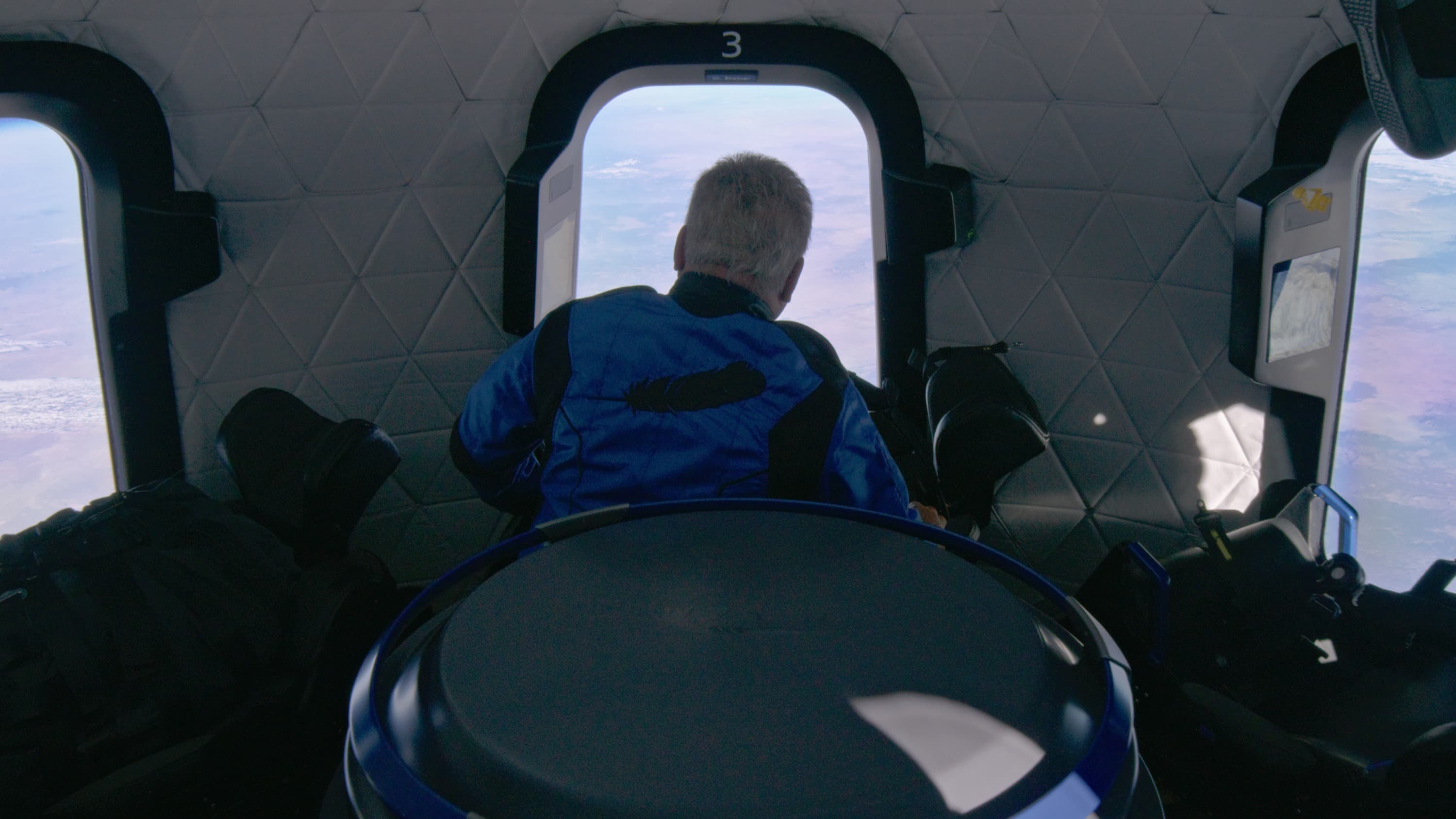William floats inside the crew capsule looking out the window at Earth's brown terrain.